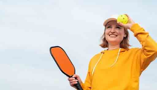 A woman in a yellow hoodie and cap holds a pickleball paddle in one hand and a yellow pickleball in the other, standing outdoors against a blue sky, celebrating wellness through active play.